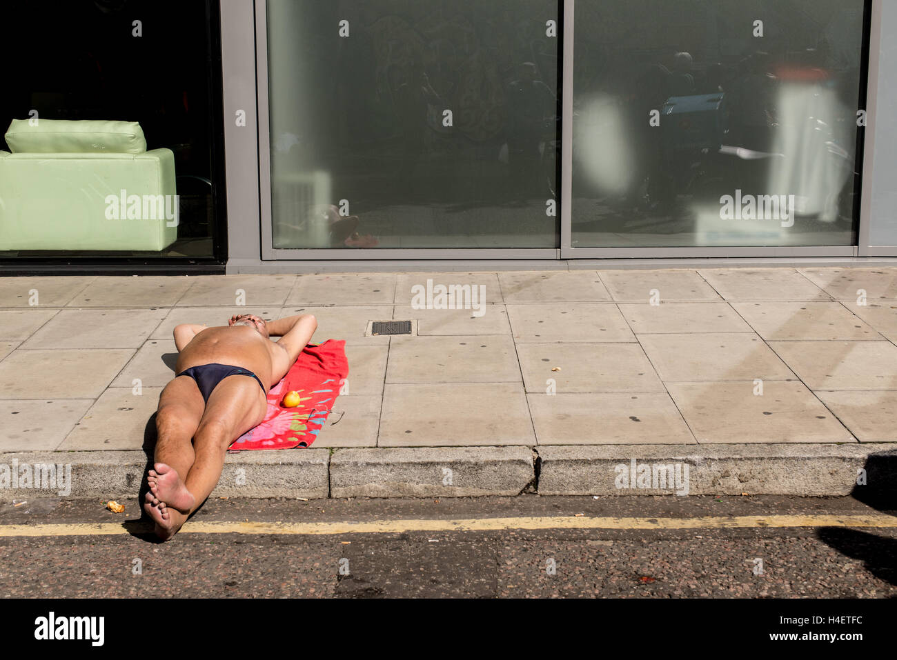 Hombre vestido con speedos acostado tomando el sol sobre una toalla roja sobre el pavimento de una calle de Londres oriental Foto de stock