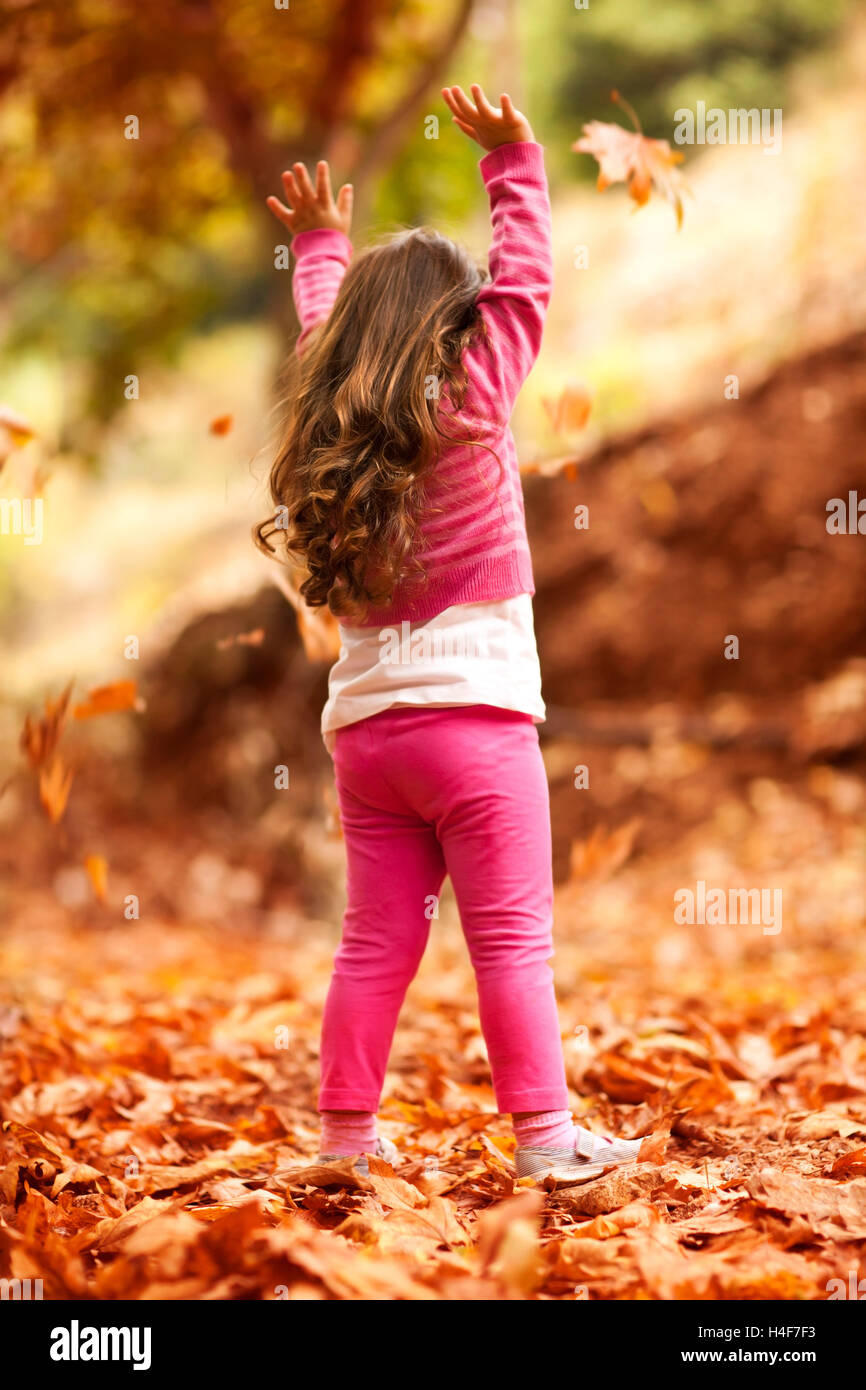 Feliz niña en otoño de parque, vista trasera de un bonito niño arrojando las hojas del árbol seco, jugar al aire libre en un cálido día de otoño Foto de stock