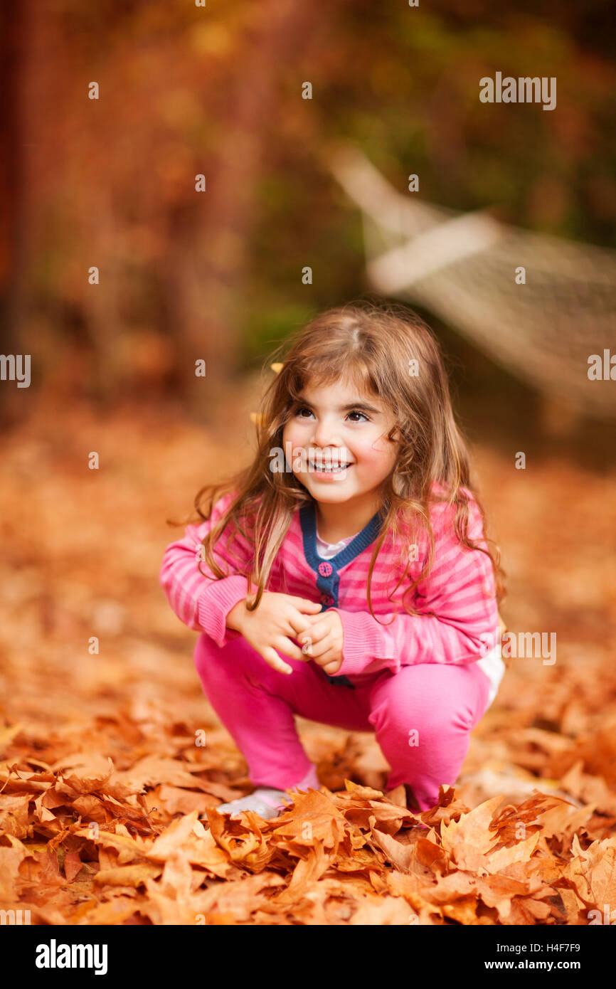 Linda niña jugando en el parque de otoño, dulce chico sentado sobre las hojas del árbol seco de color marrón, disfrutando de la belleza de la caída Foto de stock