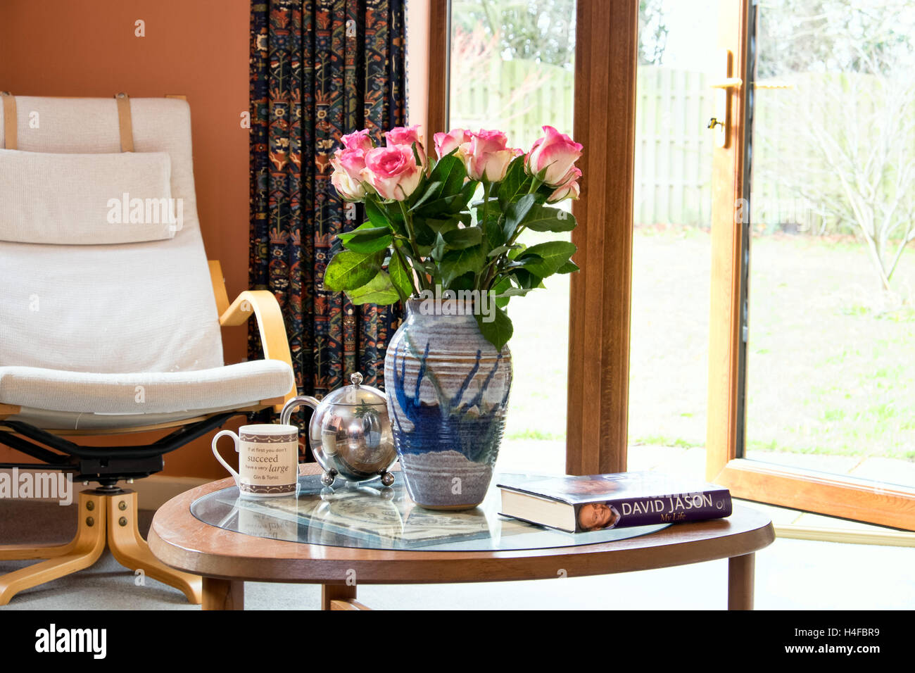 Una imagen de estilo de vida de una mesa de café con rosas, tetera, taza y reservar en una media UK casa con jardín. Foto de stock
