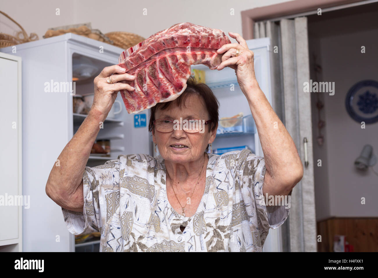 Mujer sosteniendo Senior costillas de cerdo cruda mientras está de pie delante de la nevera en la cocina abierta. Foto de stock