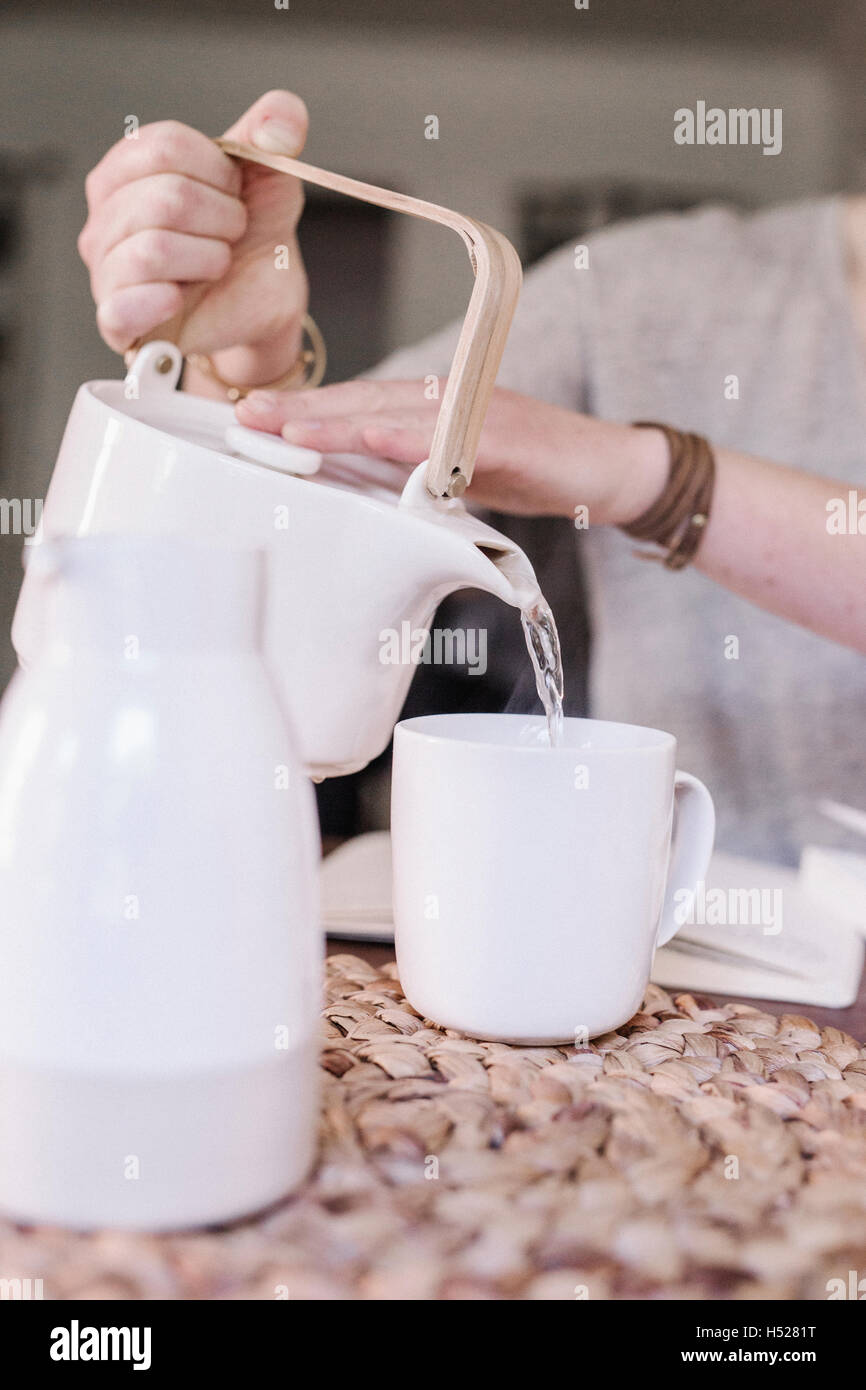 https://c8.alamy.com/compes/h5281t/mujer-sentada-en-una-mesa-en-su-apartamento-vertiendo-agua-caliente-en-una-taza-la-rutina-de-la-manana-h5281t.jpg