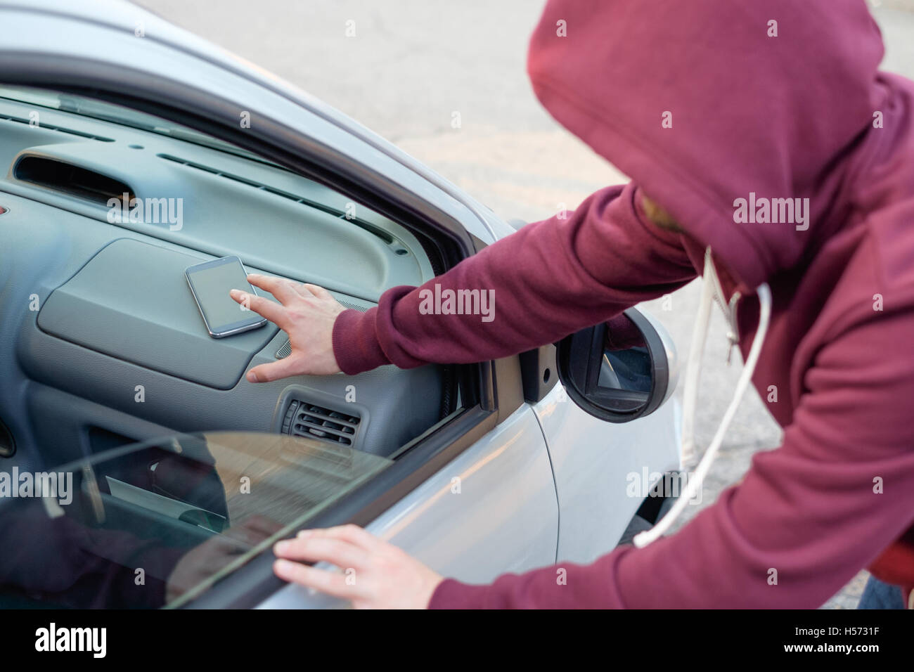Encapuchado ladrón robar un teléfono móvil desde un automóvil estacionado Foto de stock