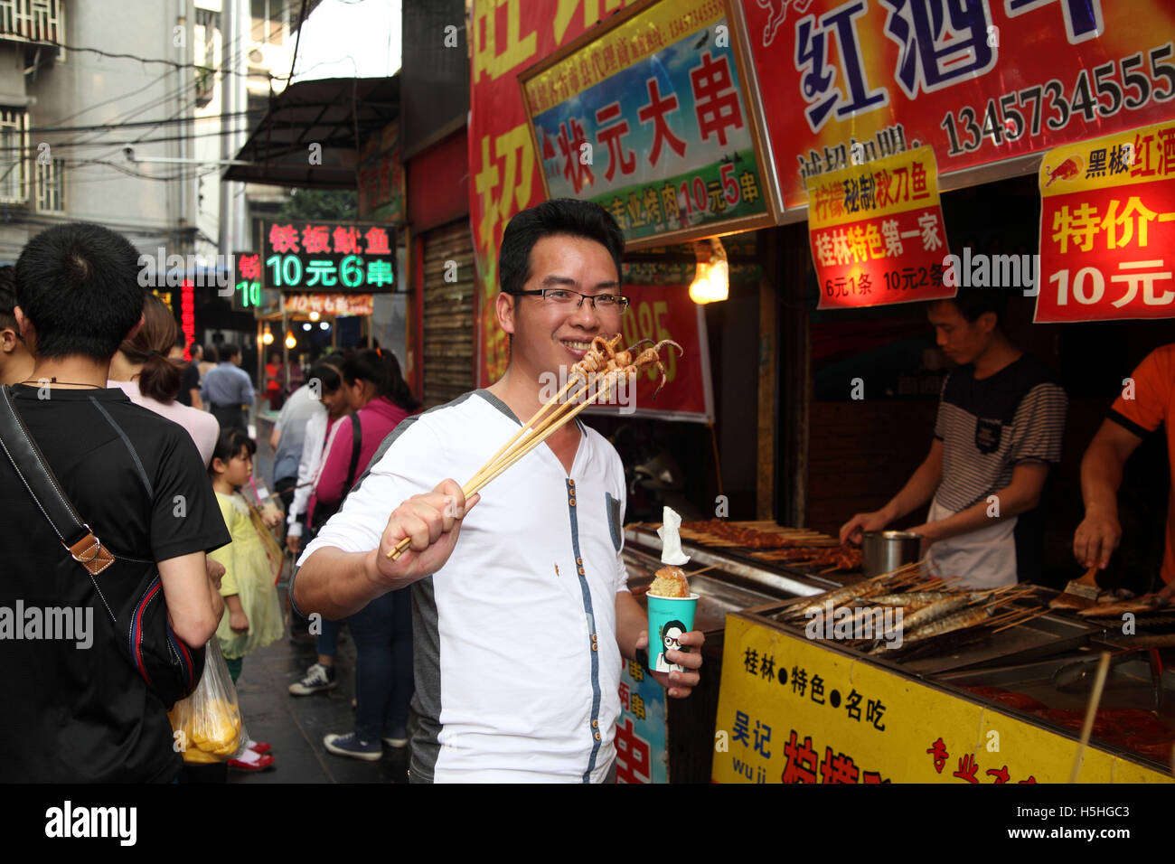 Un joven hombre chino sonríe mientras come calamares en brochetas en frente de un stand de comida rápida. El mercado en Guilin, China. 02.05.2 Foto de stock