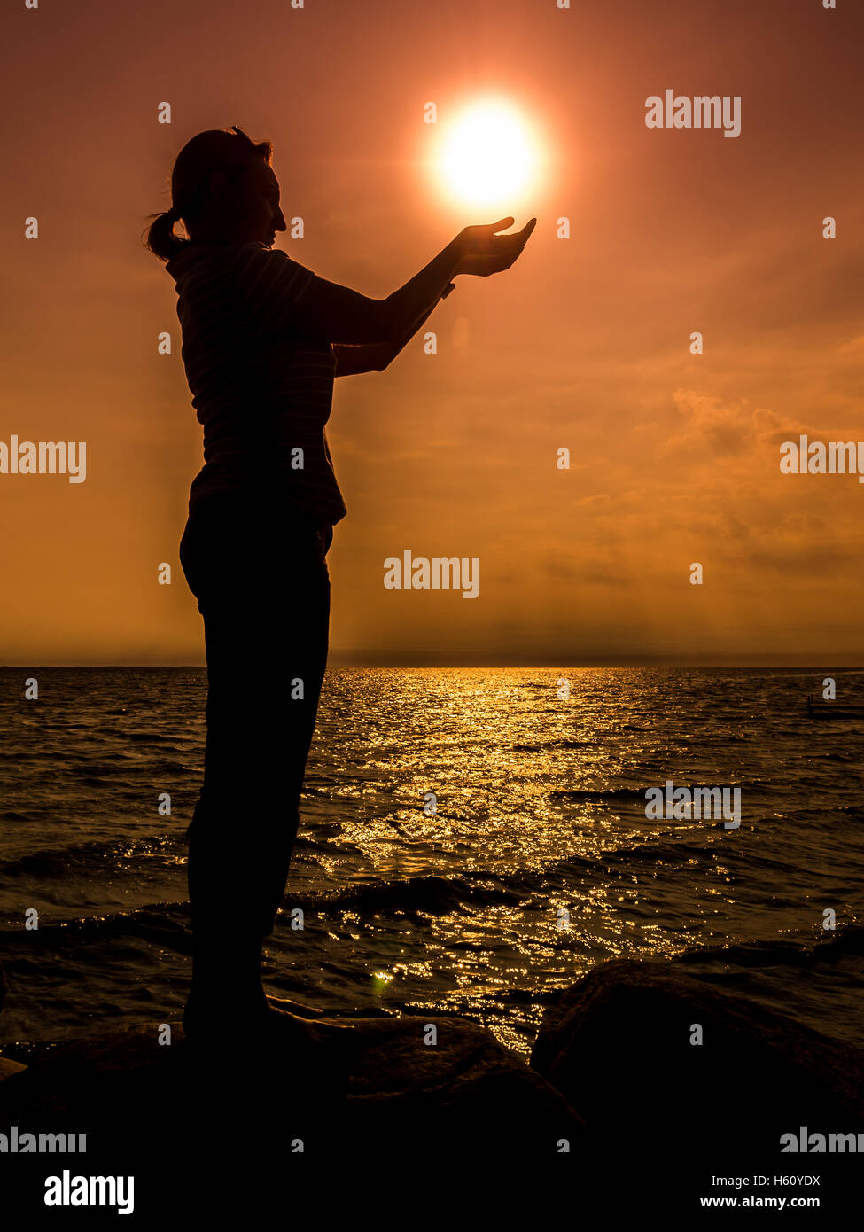 Silueta de mujer joven de pie junto a la orilla del mar sujetando el sol naciente sobre cielo naranja Foto de stock