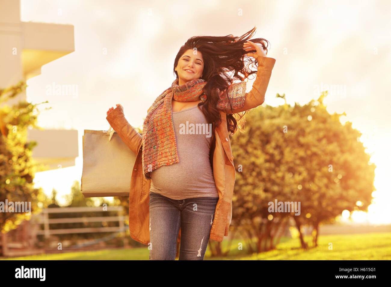 Mujer embarazada después de las compras, feliz chica con bolsas de papel expectante yendo a casa, suave luz del atardecer, gozando de gran venta de otoño Foto de stock