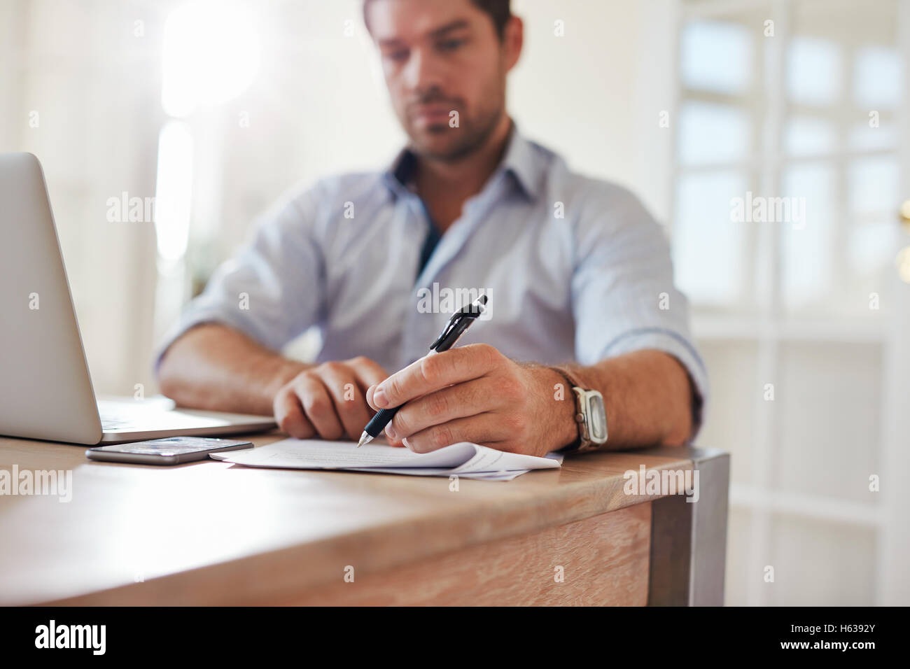 Foto de joven empresario sentado en su oficina doméstica con laptop y firmar documentos. Joven trabajando desde casa. Foto de stock