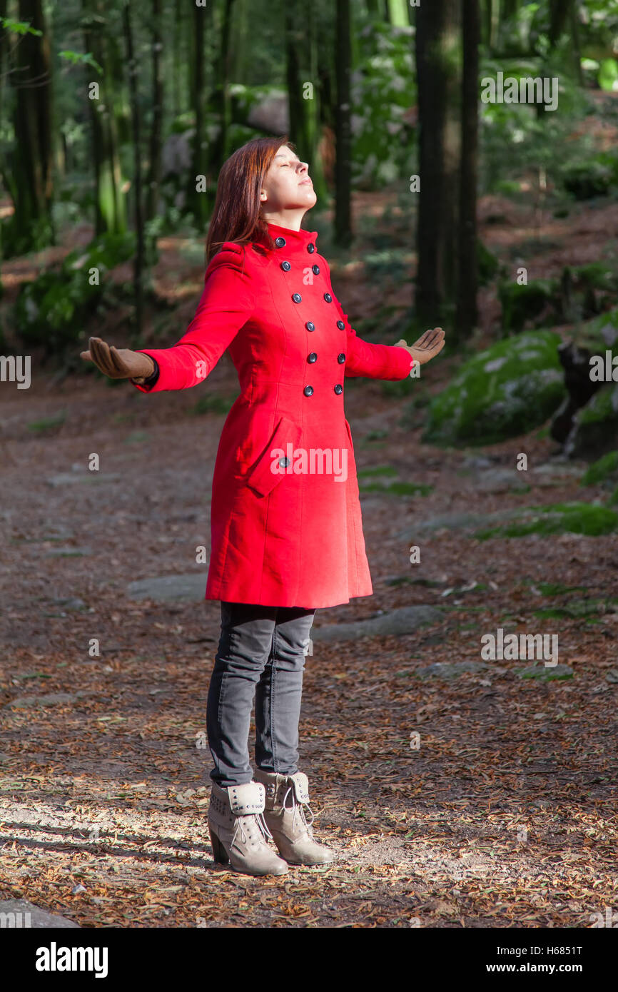 Mujer con los brazos abiertos, disfrutando de la calidez del sol de invierno en un bosque vistiendo un abrigo rojo Foto de stock