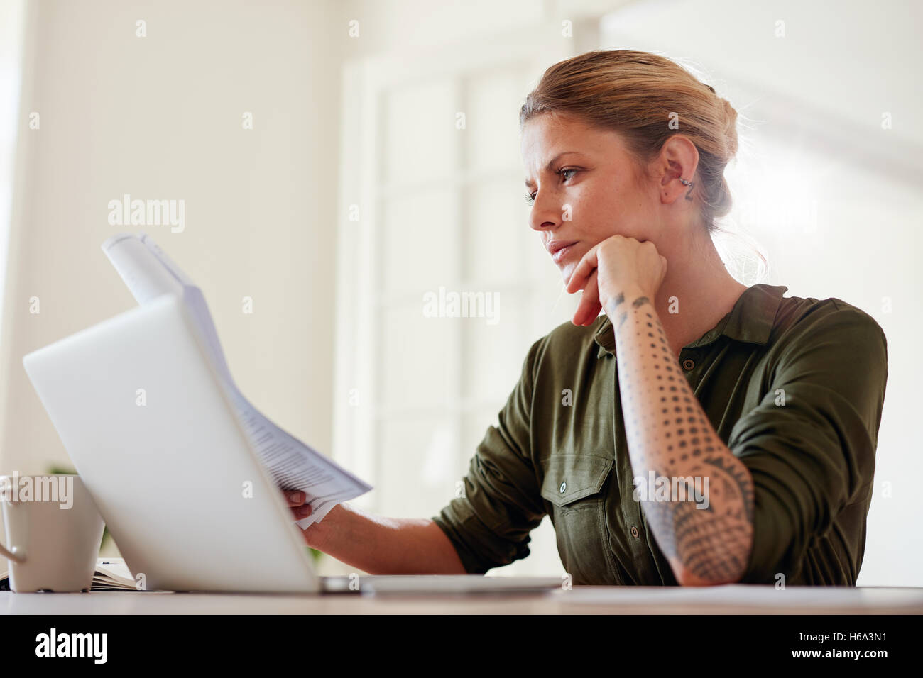 Foto de joven mujer leyendo documentos mientras se trabaja en una computadora portátil. Mujer sentada en una mesa en el interior y la lectura de algunos trámites. Foto de stock