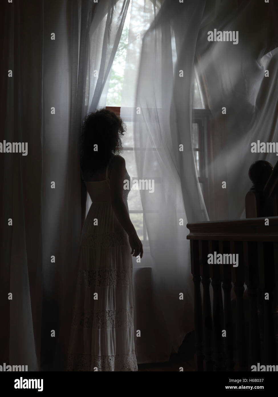 Licencia e impresiones en MaximImages.com - Mujer joven en un vestido blanco parado junto a una ventana con volar en las cortinas del viento en una habitación oscura de una casa Foto de stock