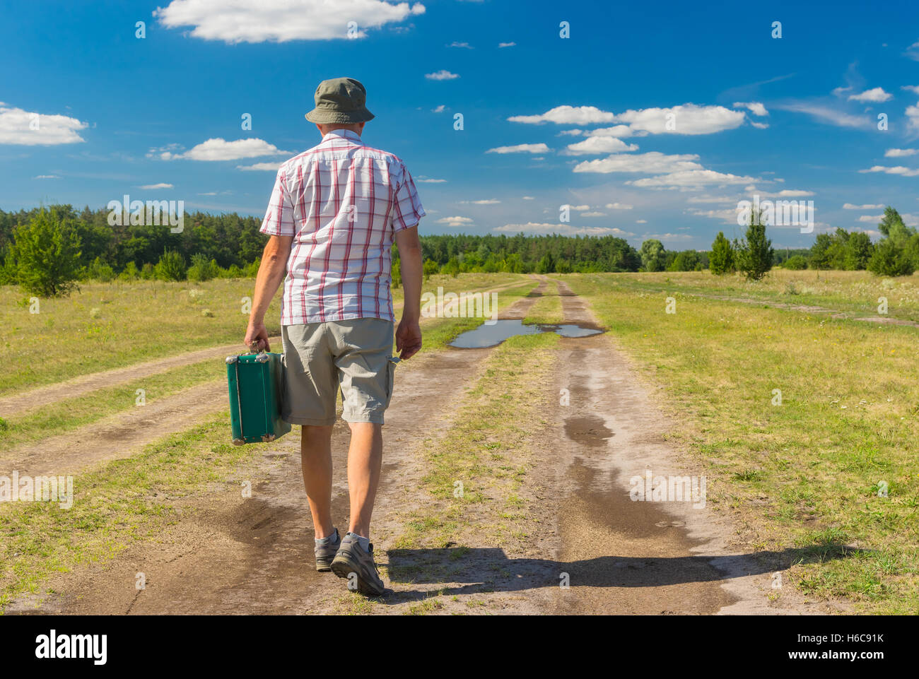 Hombre maduro vistiendo anteojeras, camisa y sombrero caminando sobre un verano country road con maleta verde Foto de stock