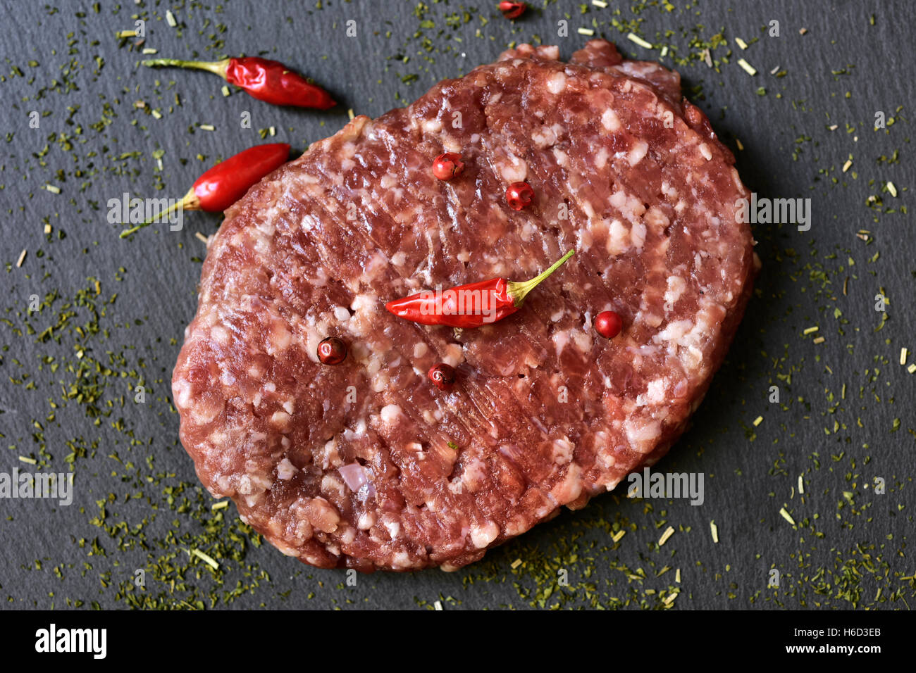 Primer plano de una hamburguesa cruda condimentada con pimienta roja, pimientos y hierbas en una superficie de pizarra Foto de stock