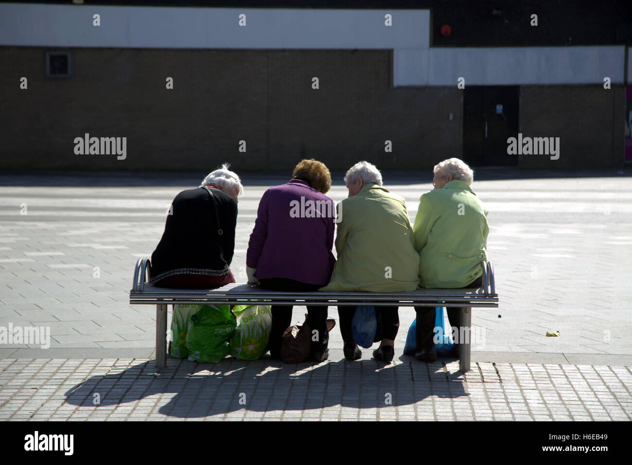 Cuatro abuelas con gemelos en un banco chismeando desde detrás con bolsas sentado Foto de stock