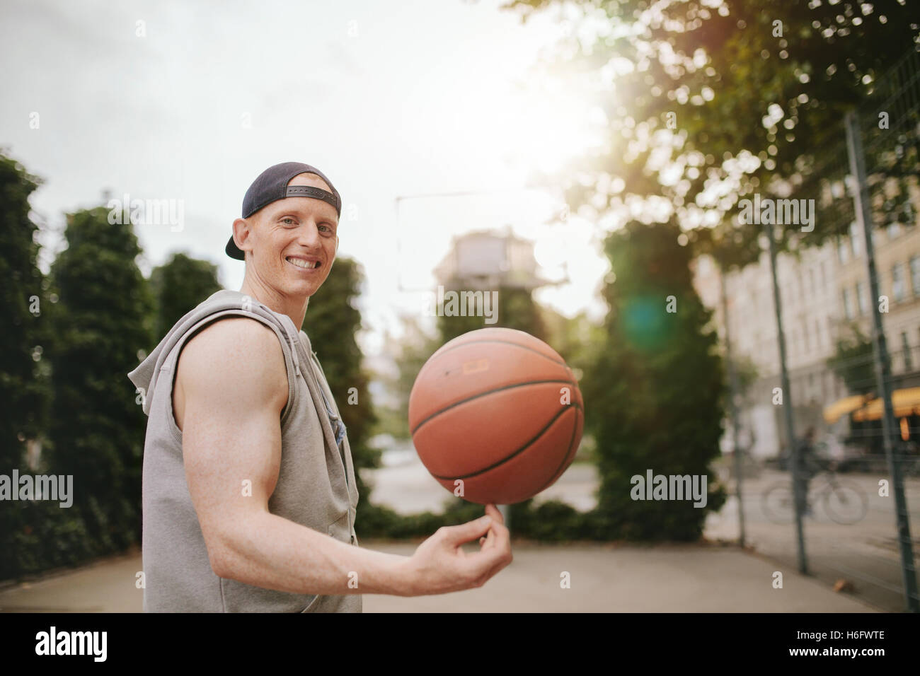 Retrato de streetball sonriente jugador gira la bola en la corte al aire libre. Feliz joven baloncesto equilibrado sobre su dedo. Foto de stock