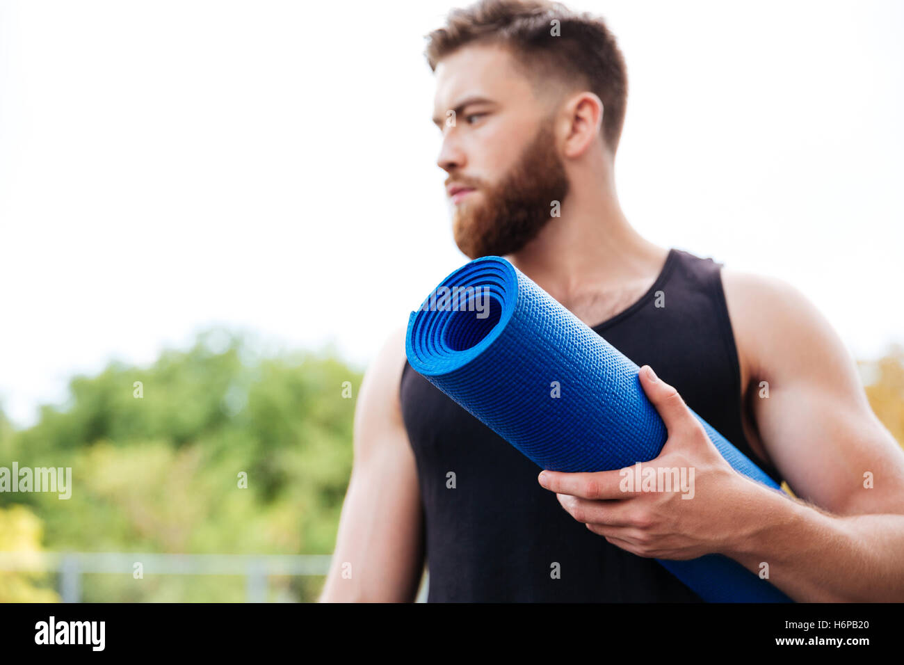 Instructor de yoga masculina grave explotación mat y apartar la mirada de pie afuera Foto de stock