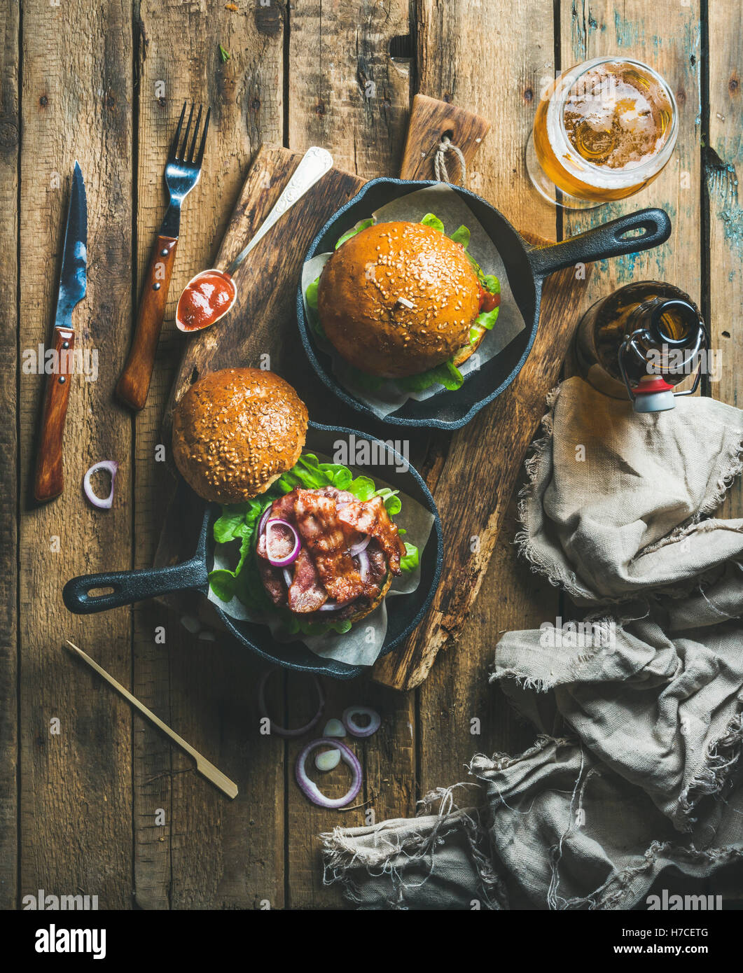 Hamburguesas de carne casero con tocino crujiente y verduras en pequeñas ollas de hierro fundido y vidrio de cerveza de trigo en junta rústico más shab Foto de stock