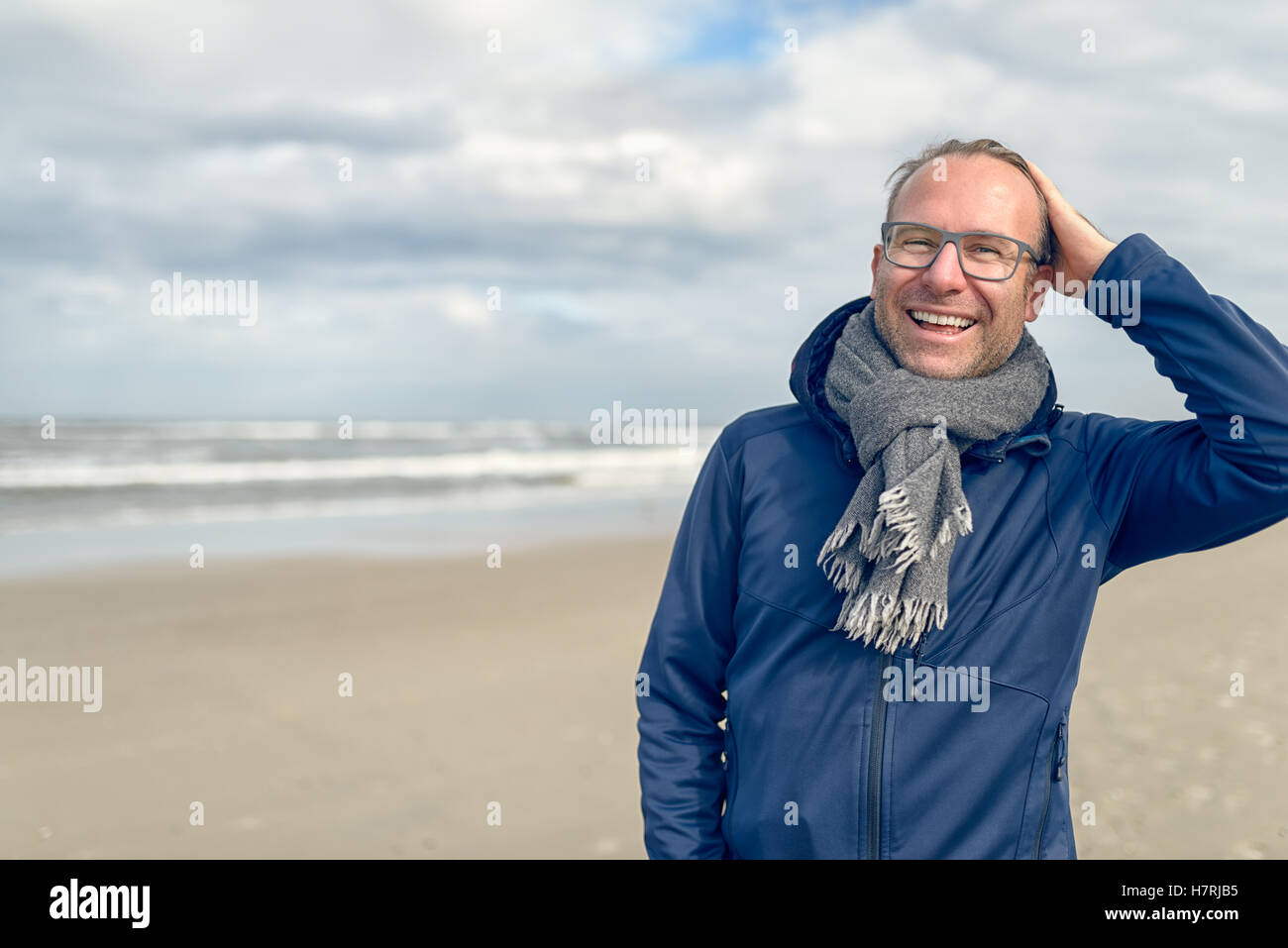 Feliz riendo hombre de mediana edad con gafas y una bufanda de lana tejida de pie en una desierta playa de otoño en un día nublado w Foto de stock