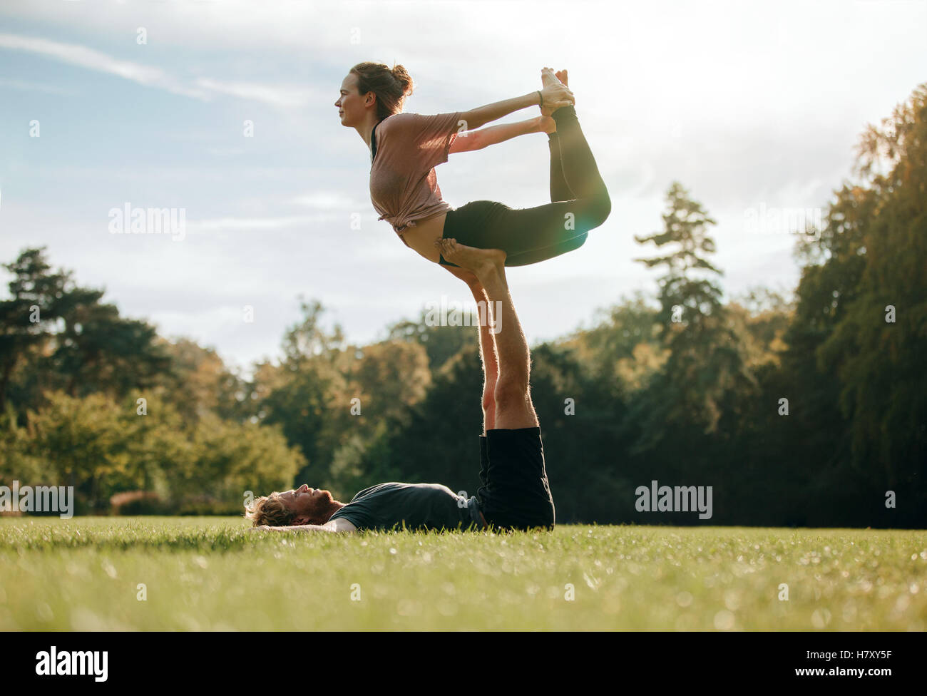 Colocar pareja joven haciendo acro yoga. Hombre tumbado sobre el césped y equilibrar la mujer en sus pies. Equilibrio acrobático en el parque. Foto de stock