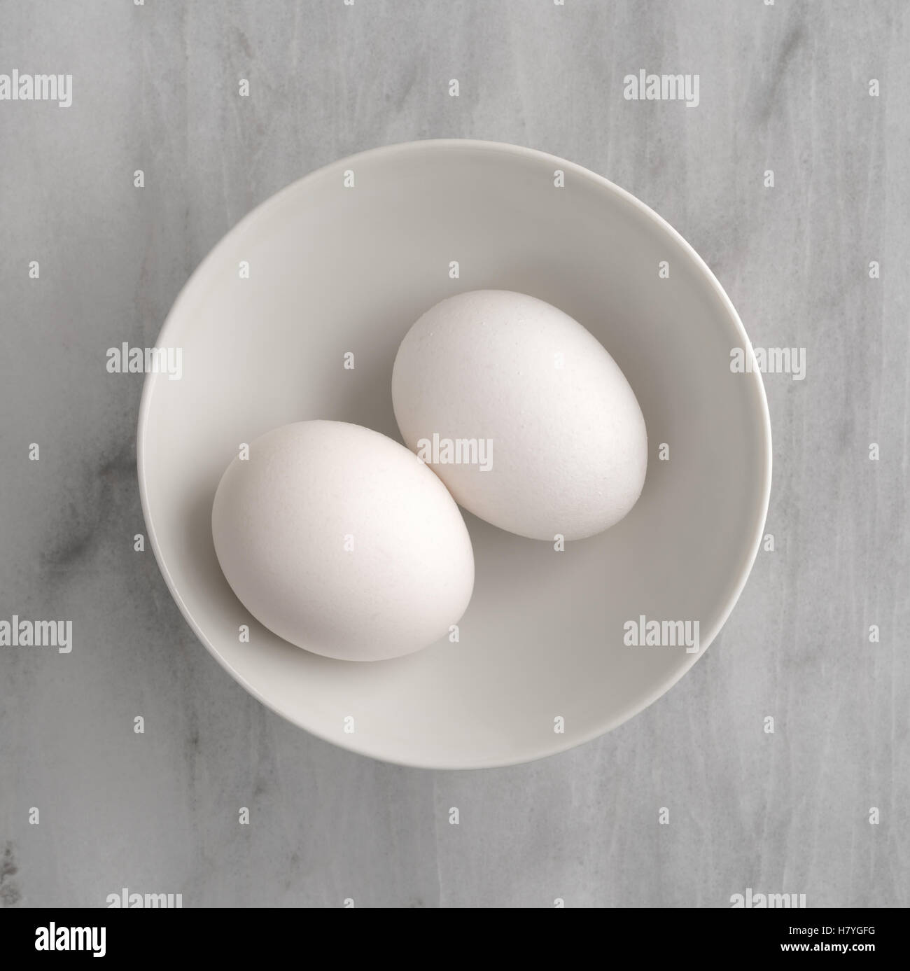 Vista superior de dos huevos en un recipiente blanco sobre una placa de corte de mármol gris. Foto de stock