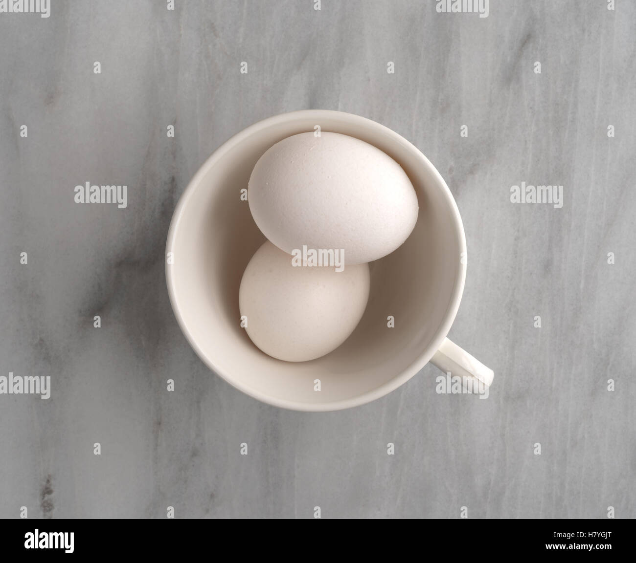 Vista superior de dos huevos en una taza blanca sobre una placa de corte de mármol gris. Foto de stock