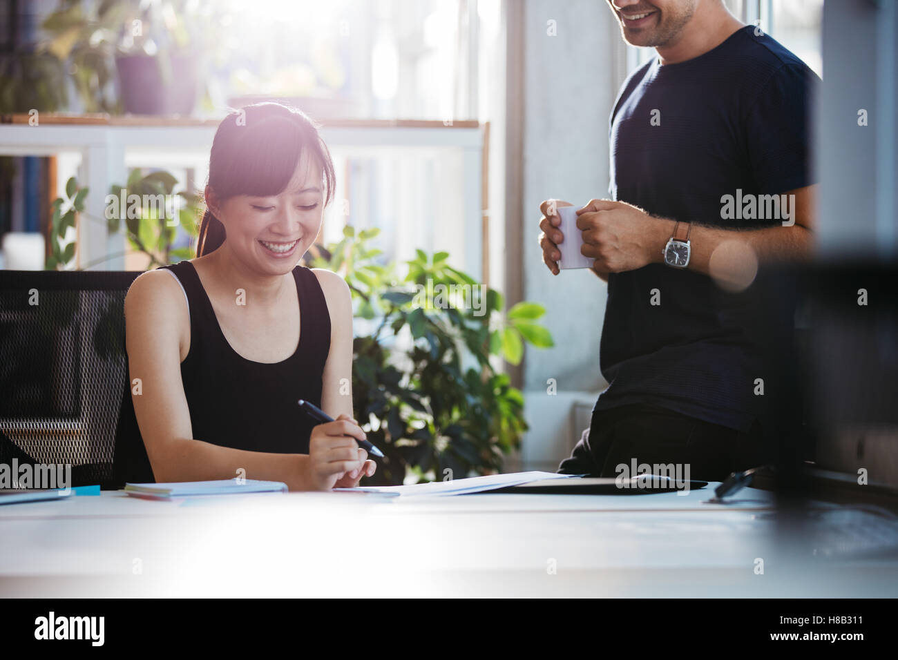 Foto de mujer sonriente sentada en su escritorio trabajando con colega masculino en pie. La gente de negocios en el trabajo en la oficina. Foto de stock