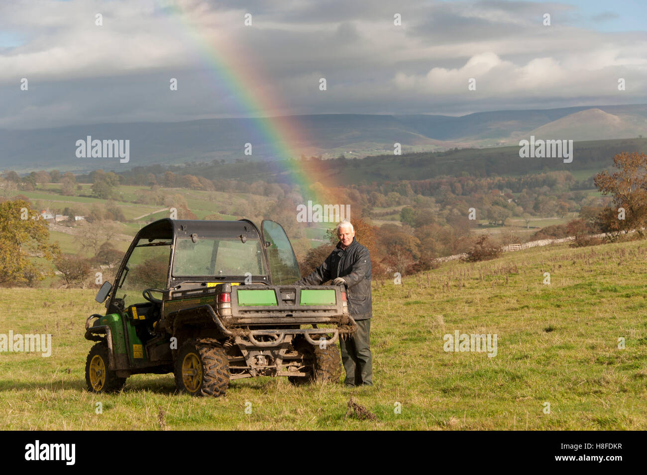 Los pastos de montaña en agricultor, apoyándose en un vehículo agrícola 4x4, con un arco iris detrás de él. Foto de stock