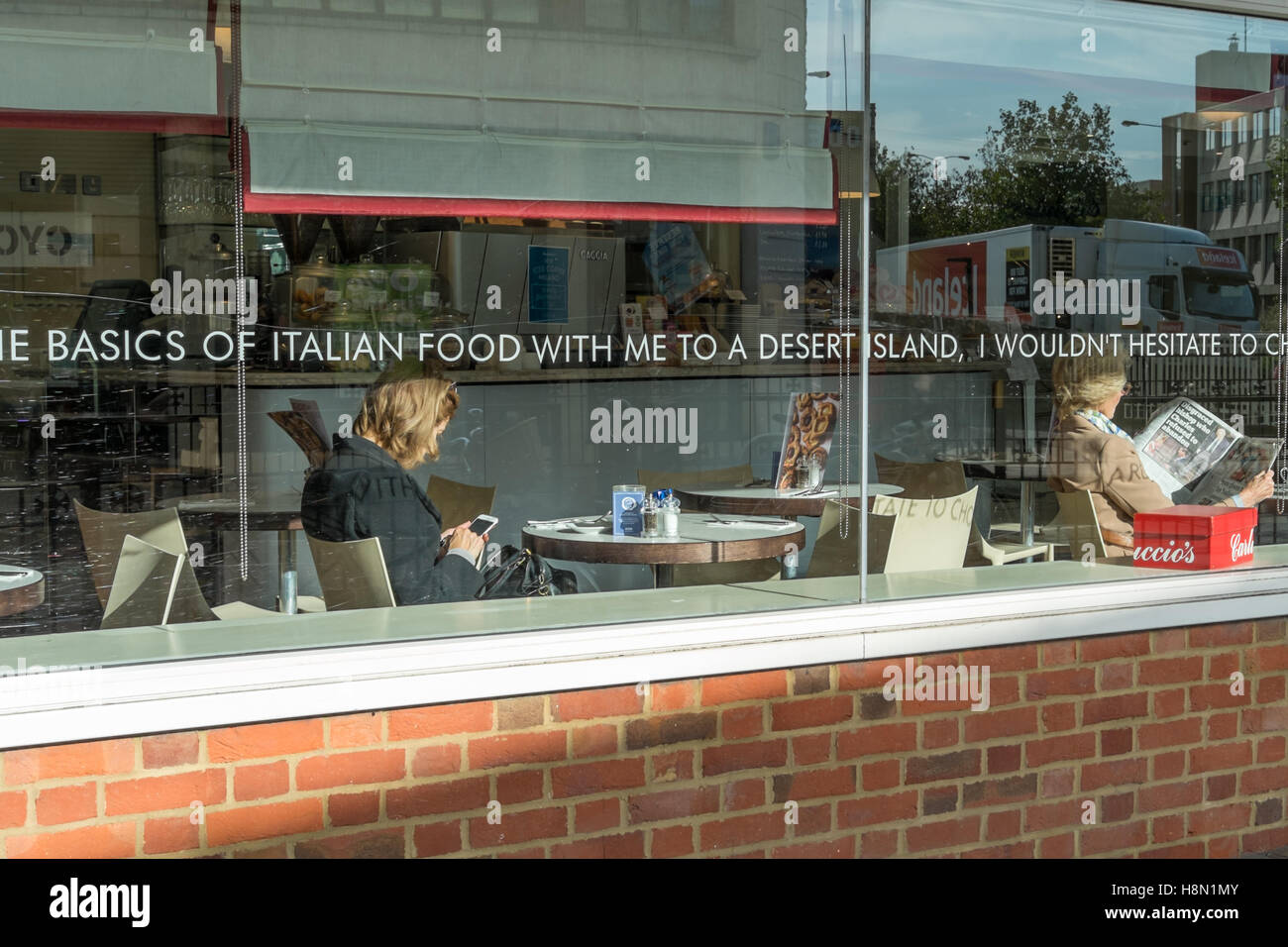 Dos mujeres sentadas por la ventana en un bistro en tablas separadas. Uno  está leyendo un periódico y el otro está mirando su teléfono móvil  Fotografía de stock - Alamy