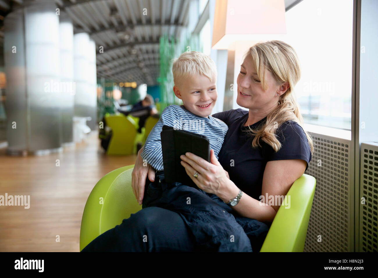 Mujer madura, sentado en el hall del aeropuerto y con tablet con boy (6-7) sentado en su regazo Foto de stock