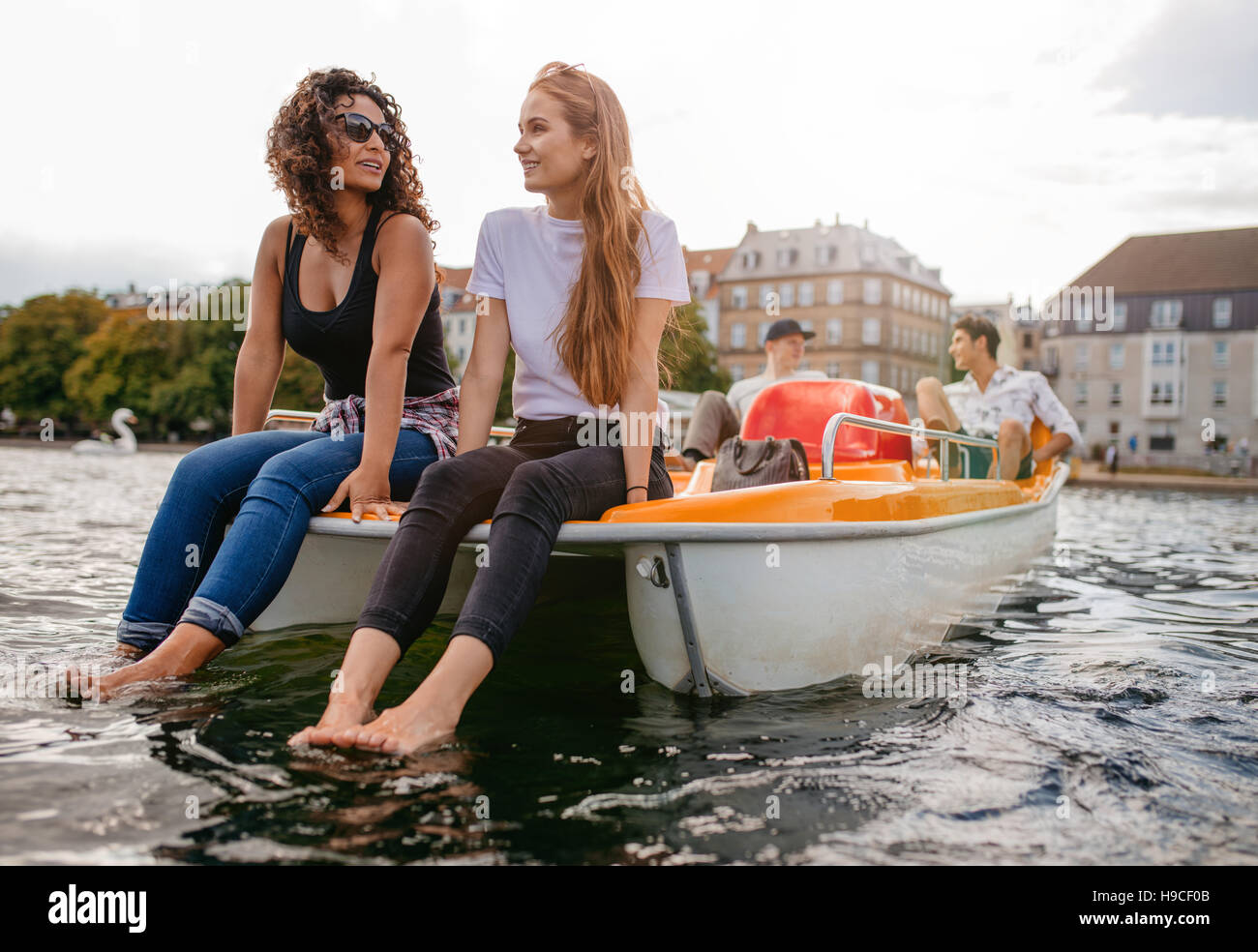 Tiro de dos mujeres sentadas en bote a pedal frontal con los pies en el agua y el hombre en el fondo. Amigos adolescentes disfrutar de paseos en bote en el lago. Foto de stock