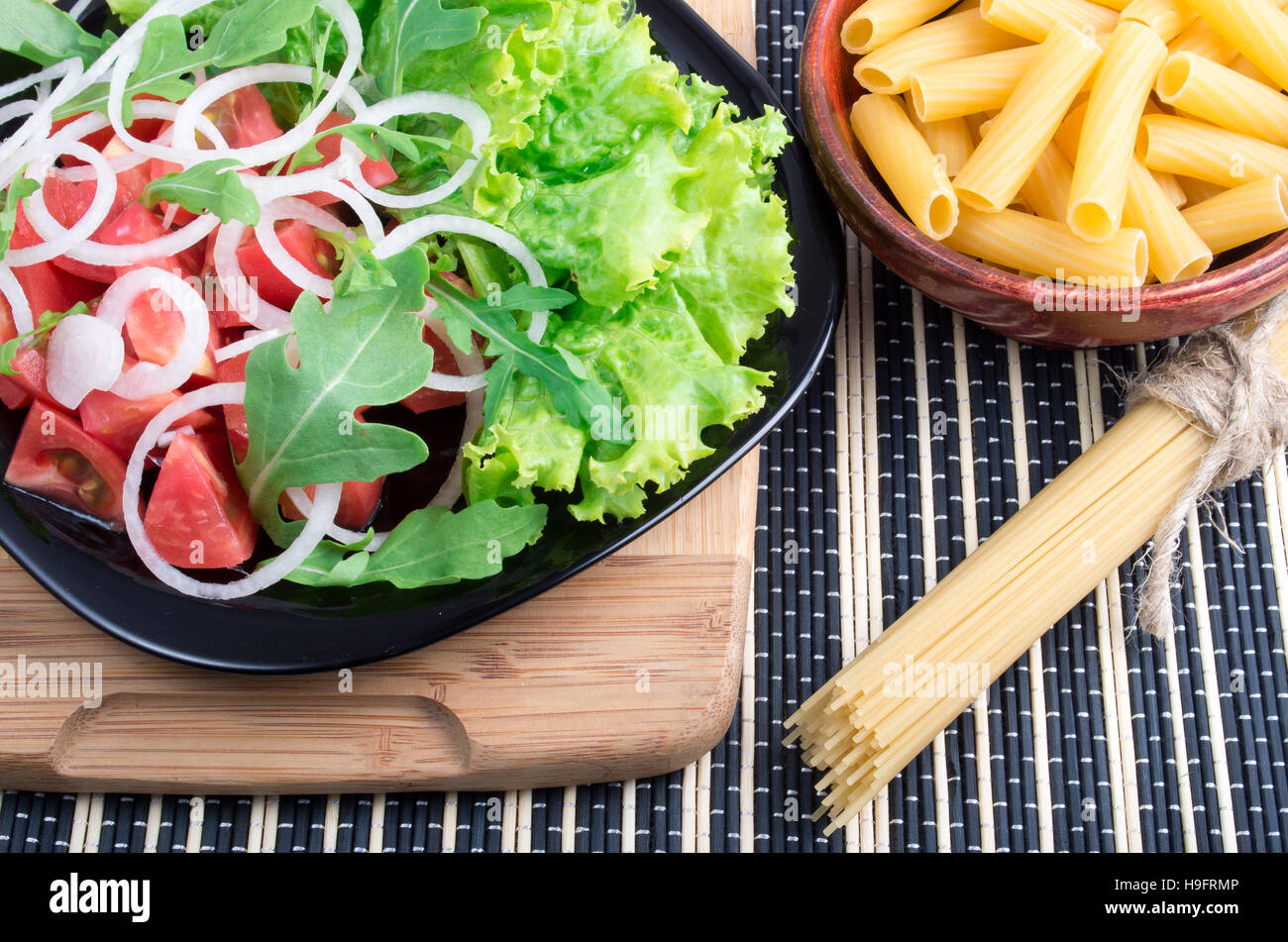 La composición de la mesa con una ensalada fresca de tomates, cebollas, lechuga y rúcula sobre un soporte de madera, pasta y spaghetti para cocinar Foto de stock