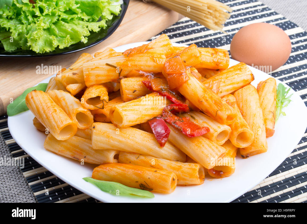 Plato de rigatoni pasta con salsa de verduras junto al plato con lechuga y los ingredientes para cocinar Foto de stock