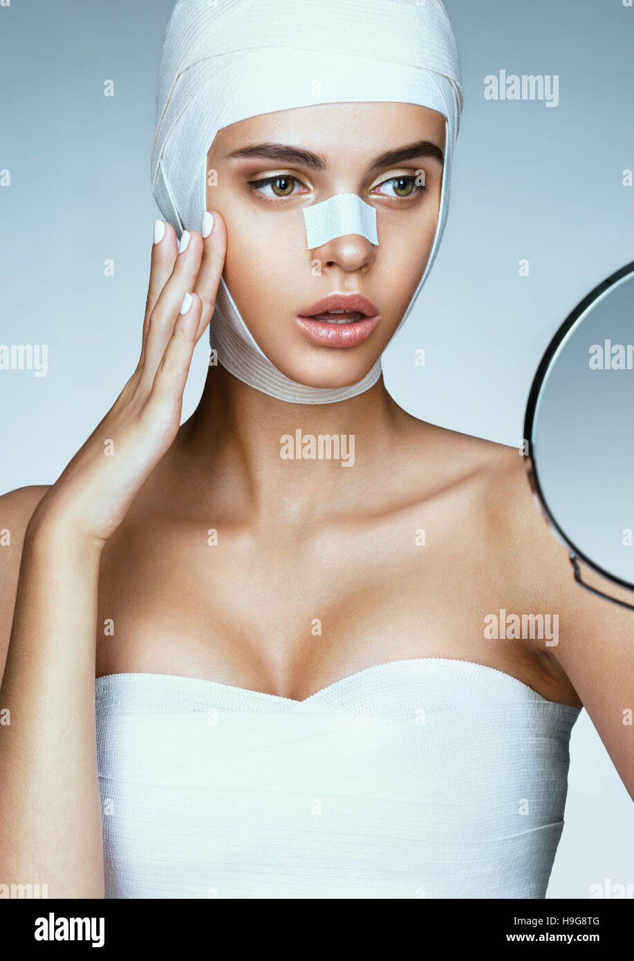 Mujer joven después de la cirugía plástica, mirando en el espejo y tocando su pómulo. Foto de mujer envuelto en vendajes médicos. Concepto de belleza Foto de stock