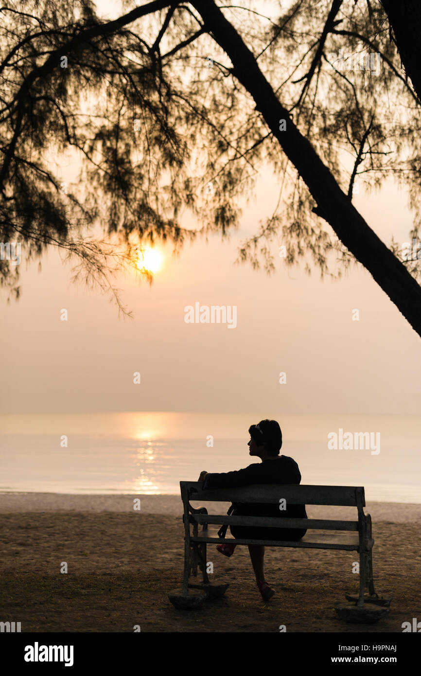 Soledad, silueta mujer viajero sentado solo con su bolsa en la banqueta retro al atardecer en la playa. Foto de stock