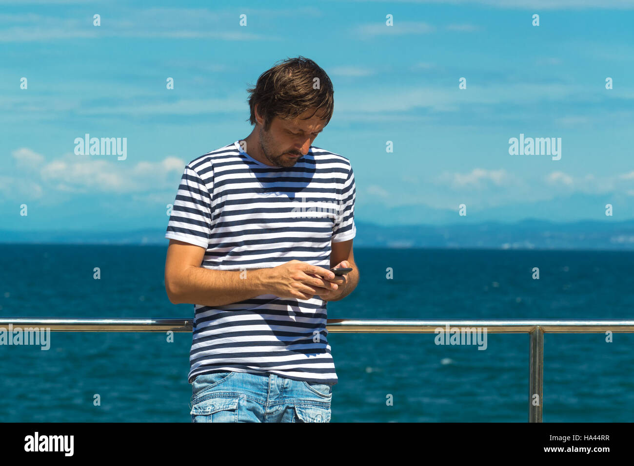 En rayas Camiseta Hombre marinero enviar SMS por el mar. A través de teléfono móvil para la comunicación sobre la hora de verano para vacaciones de playa. Foto de stock