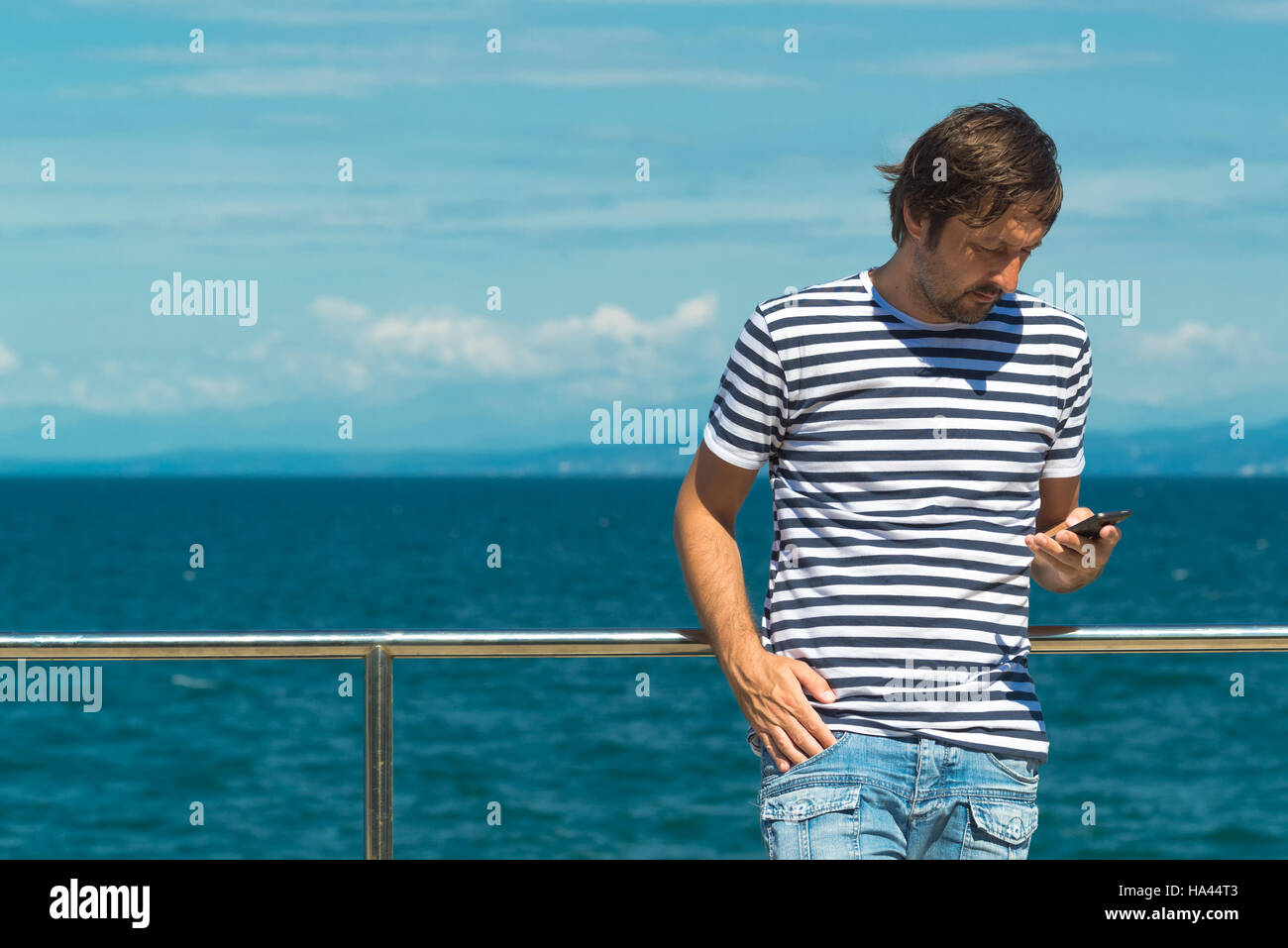 En rayas Camiseta Hombre marinero enviar SMS por el mar. A través de teléfono móvil para la comunicación sobre la hora de verano para vacaciones de playa. Foto de stock