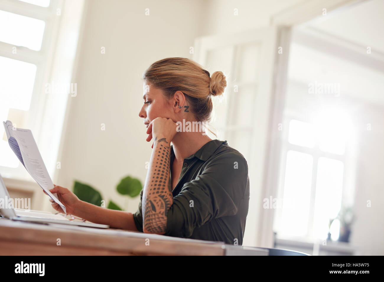 Lado retrato de mujer joven sentada a la mesa la lectura de documentos. Mujer pasando por contrato papeles en la oficina en casa. Foto de stock