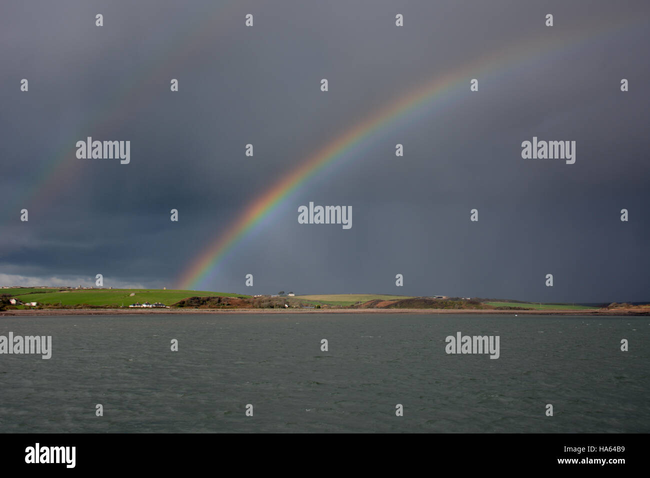 Brillante en la parte superior del arco iris doble cleddau en Pembrokeshire contra un cielo oscuro sobre aguas tranquilas Foto de stock