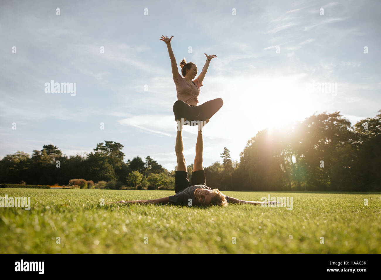 Colocar pareja joven haciendo acro yoga en el parque. Hombre tumbado sobre el césped y equilibrar la mujer en sus pies. Foto de stock