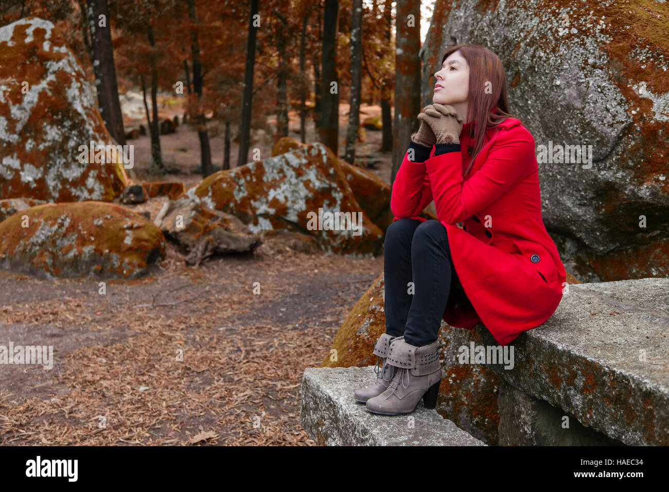Mujer joven deprimido sentado en una mesa y un banco de piedra en un bosque vistiendo un abrigo rojo durante el invierno Foto de stock