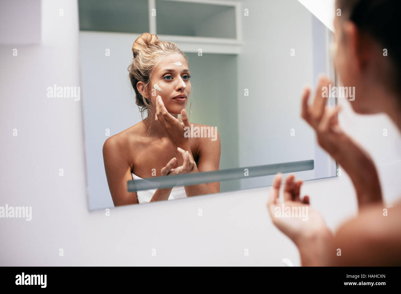 Hermosa joven aplicar crema en el baño. Mujer mirando en el espejo del baño y ponerse crema para la cara. Foto de stock