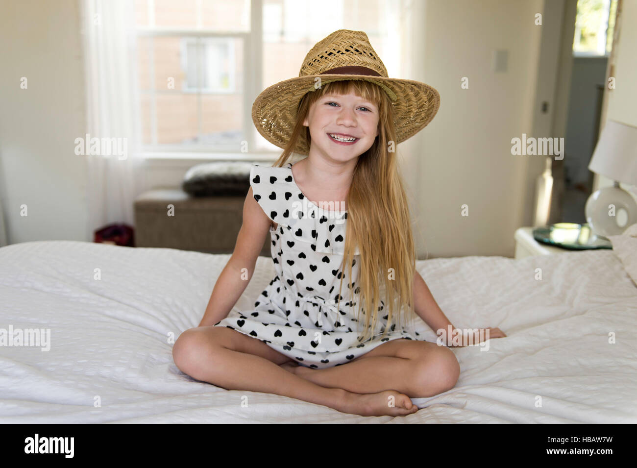 Niña sentada en la cama, sonriente, luciendo sombrero de paja Foto de stock
