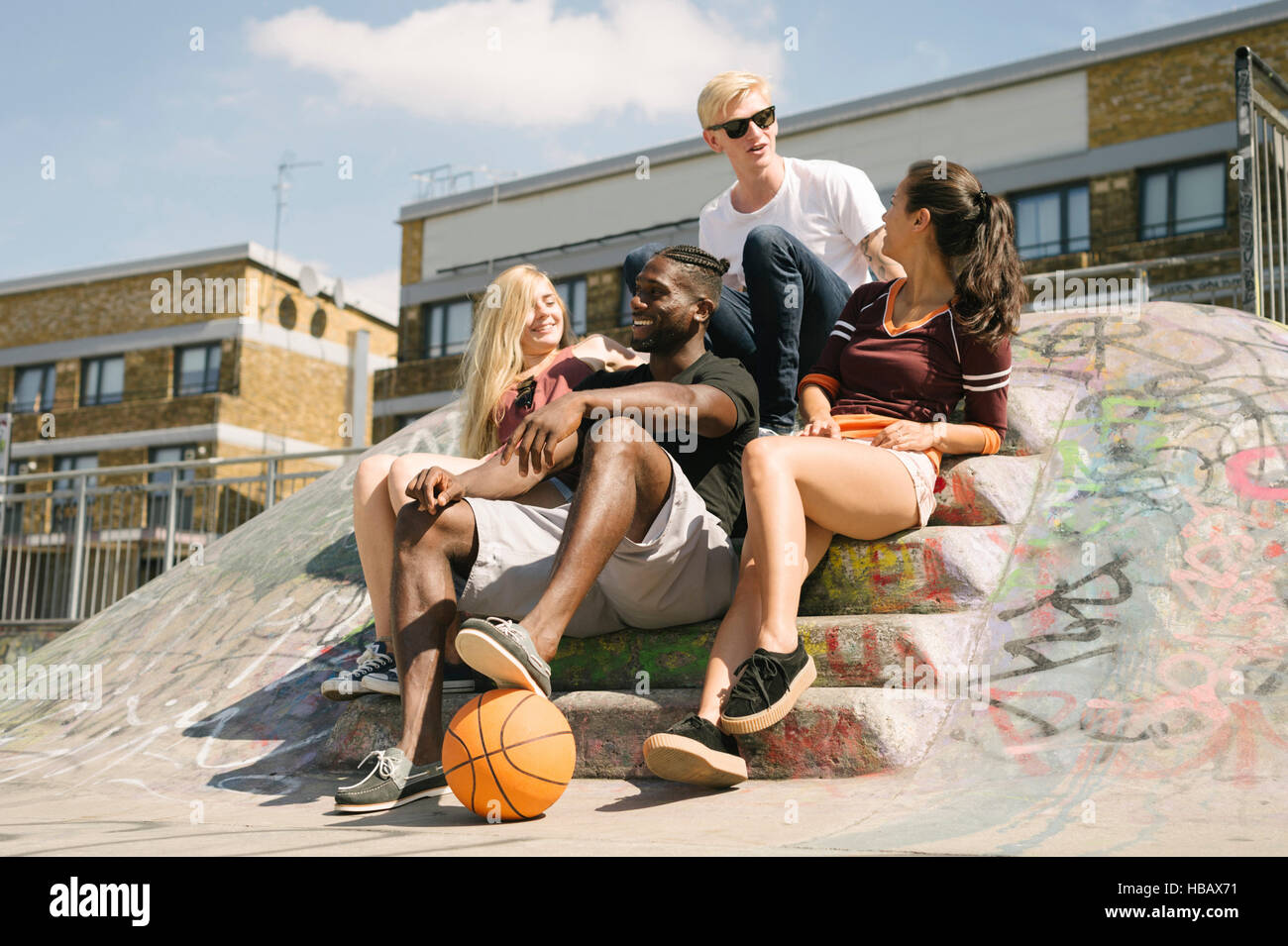 Cuatro amigos de baloncesto masculino y femenino sentado chateando en ciudad skatepark Foto de stock