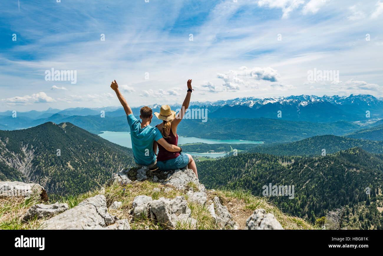 Pareja joven con los brazos en el aire, la vista del lago desde el pico Heimgarten Kochel, izquierda Herzogstand, Alta Baviera, Baviera, Alemania Foto de stock