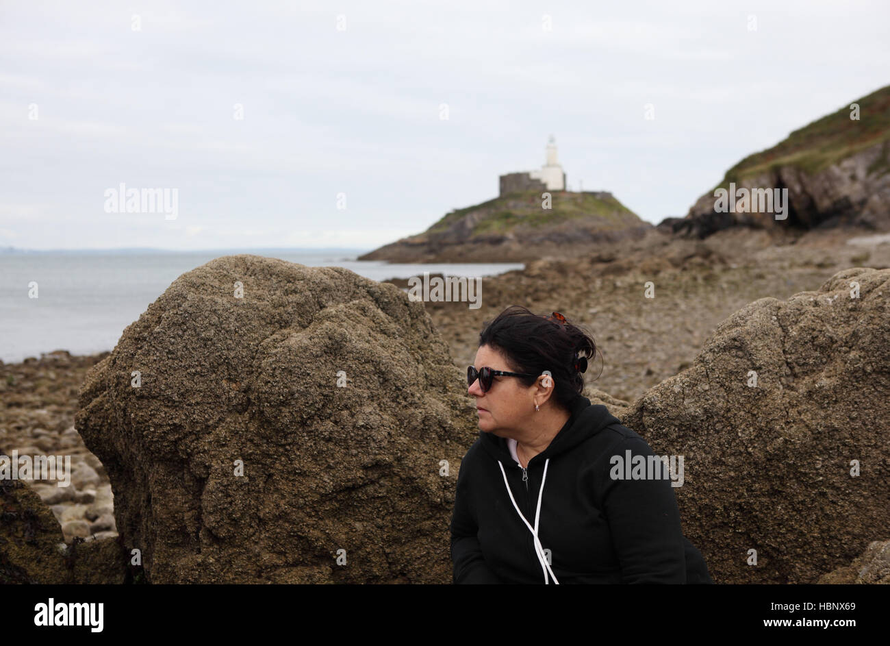 Senior Citizen, mujer de 58 años de edad, sentada por las rocas en la playa mirando el mar Celta con una luz detrás de su casa. Foto de stock