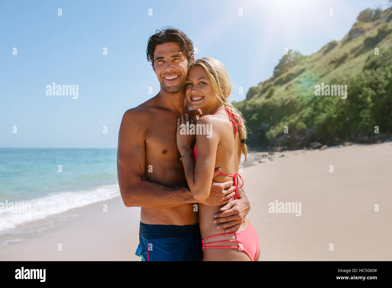 Pareja amorosa en abrazar en traje de baño en la playa. Joven pareja romántica en la orilla del mar en busca de distancia y sonriente. Foto de stock