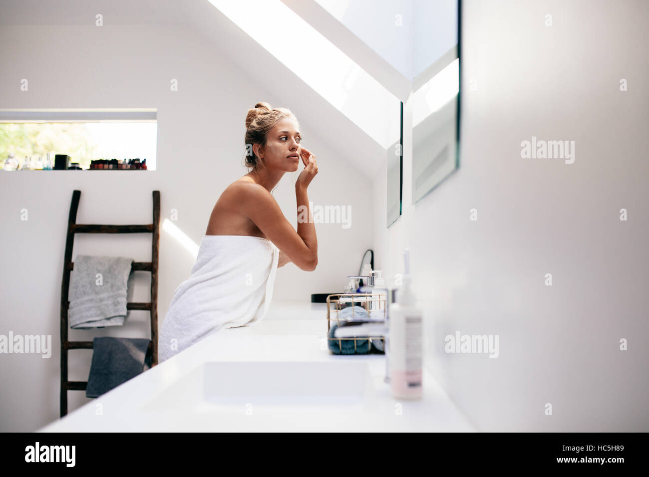 Mujer joven envuelta en una toalla, mirando en el espejo y aplicar crema en su cara. Mujeres teniendo cuidado de la piel de la cara en el baño después del baño. Foto de stock