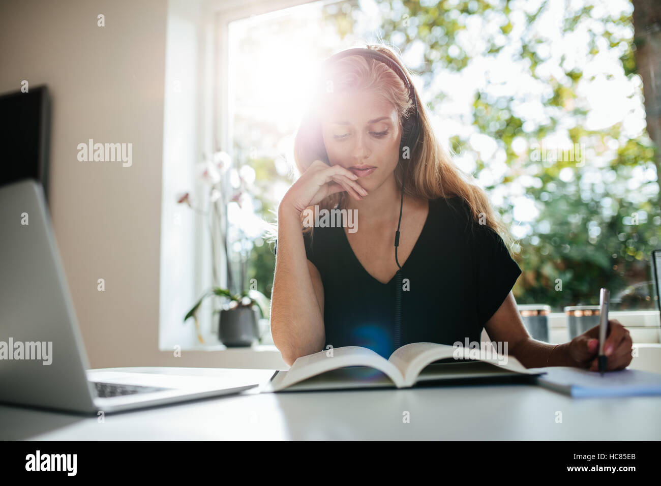 Mujer joven sentado en la mesa con el portátil y tomar notas en el libro. Estudiante femenino en casa. Foto de stock