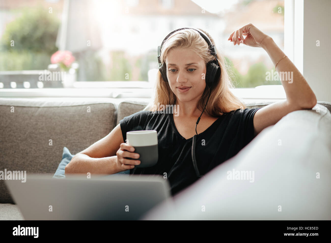 Hermosa joven con auriculares sosteniendo una taza de café y mirando el portátil. Mujer sentada en la cama en la sala de estar como en casa. Foto de stock