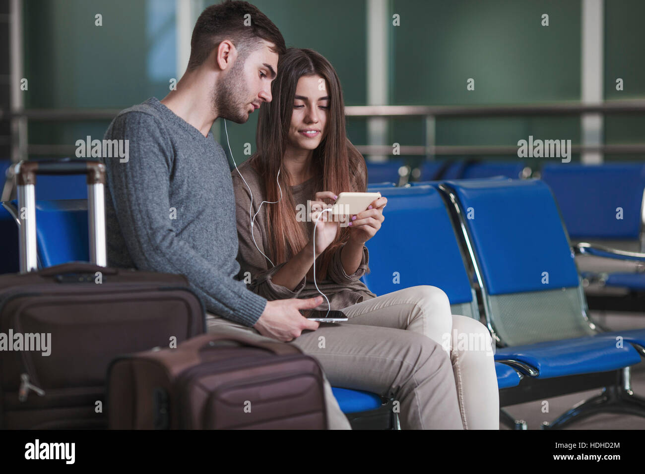 Pareja joven escuchando música a través de teléfono inteligente mientras espera en el aeropuerto Foto de stock