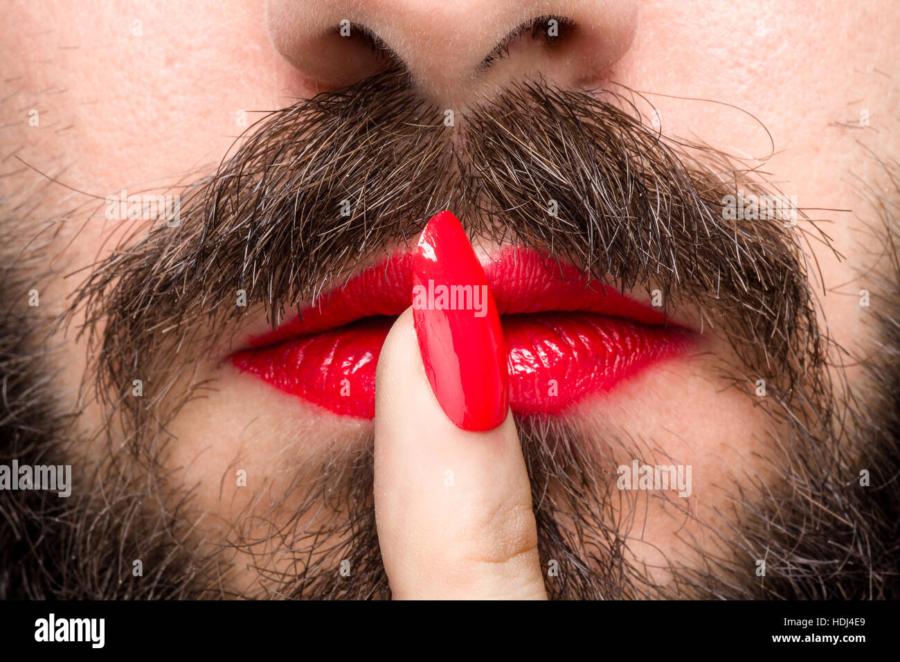 Hombre Barbado con lápiz labial rojo en los labios y esmalte de uñas haciendo gestos de silencio Foto de stock